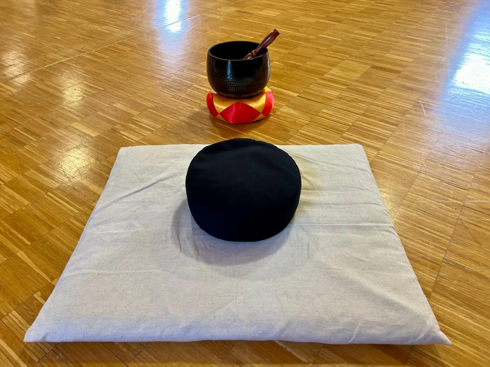 Meditationsmatte mit schwarzem Zafu-Kissen, dahinter eine Klangschale auf Holzboden